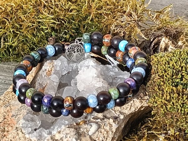 Magnifique bracelet conçu à partir de perles en ébène, et de perles tchèques en verre. Rayonne les énergies de protection, purification, harmonisation, sagesse, ancrage… L’ébène naturelle est le nom donné au bois de cœur produit par plusieurs espèces d’arbres de la famille des Ebenaceae appartenant au genre Diospyros, appelés ébéniers, ainsi que certains arbres du genre Dalbergia. Ils se rencontrent dans les régions tropicales de l’Ancien Monde et sont connus depuis la plus haute Antiquité pour la couleur très sombre, proche du noir, de leur bois. Symbole de puissance, de pureté, de protection et de chance. Depuis l’antiquité on lui attribue de nombreuses propriétés magiques. Telles que chasser les mauvais esprits et les démons. La croyance veut aussi que le bois d’ébène soit un grand réceptacle des énergies magiques, aussi on y enfermait dans des coffres en ébène les objets magiques. L’ébène est le bois le plus puissant en magie et divination. Son pouvoir est général, rien ne lui résiste et il agit dans tous les domaines. Très protecteur, il convient mieux aux expérimentés. Les bijoux en bois d’ébène apporteraient courage, force et agilité. Les perles tchèques, sont de couleurs bleues, violettes, vertes, marrons… Chacune des couleurs étant vibratoires, car ré-harmonisent les chakras de la gorge (communication), chakras couronne (sagesse) et chakras du cœur (amour). L’arbre de vie : D’une jeune pousse devenue un grand arbre majestueux, il évoque l’énergie de la vie. L’arbre de vie peut donc être comparé à l’Homme qui grandit au fil du temps et de ses expériences. Chez les chrétiens, il est souvent associé à l’amour de Dieu et évoque sa protection. Symbole universel et intemporel, l’arbre de vie constitue une représentation particulièrement puissante du cycle de la vie. Ses racines représentent les origines qui s’ancrent à la terre où il puise son énergie. Ses branches symbolisent le développement de la vie tandis que ses feuilles évoquent l’épanouissement et l’accomplissement de soi.  L’arbre de vie peut représenter l’homme durant le cycle de sa vie, de sa naissance à sa mort. Dans de nombreuses religions et civilisations, l’arbre de vie revêt d’un symbolisme particulier et propre à chacun. Mais toutes les croyances s’accordent sur le fait que l’arbre de vie symbolise entre autres la renaissance, l’amour et la protection. Il est également symbole d’énergie, de sagesse et de longévité.